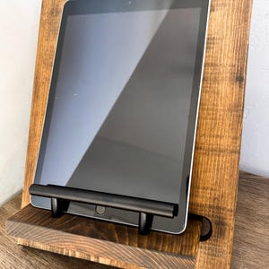 May include: A wooden tablet stand with a dark stain finish. The stand features a shelf and a black metal bar to hold the tablet in place. The tablet screen is black, and the stand is made of wood.