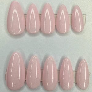 May include: A set of ten almond-shaped press-on nails in a light pastel pink color with a subtle glitter finish. The nails are arranged in two rows of five, showing a gradient of sizes from the largest thumb nail to the smallest pinky nail.