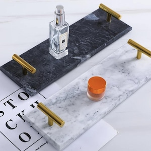 Könnte beinhalten: Zwei rechteckige Marmortabletts mit goldfarbenen Griffen. Ein Tablett ist schwarz mit einer Parfümflasche, das andere ist weiß mit einem kleinen Glas. Die Tabletts stehen auf einer weißen Oberfläche.
