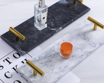 May include: Two rectangular marble trays with gold-coloured handles. One tray is black with a perfume bottle, and the other is white with a small jar. The trays are on a white surface.