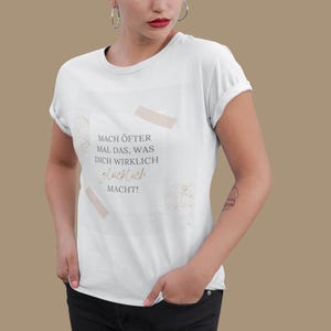 Könnte beinhalten: Weißes T-Shirt mit Rundhalsausschnitt und hochgekrempelten Ärmeln. Das Shirt zeigt den deutschen Text "Mach öfter mal das, was dich wirklich glücklich macht!" in einer Mischung aus Schriftarten und Farben. Das Design umfasst beige Klebebandformen.