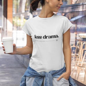 Könnte beinhalten: Weißes T-Shirt mit dem Text "less drama." in schwarzer, fetter Schrift. Darunter steht "MORE DREAMS" in kleinerer Schrift. Das Shirt wird mit grauer Jogginghose und einer um die Taille gebundenen Jeansjacke getragen.