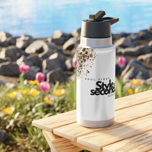 Könnte beinhalten: Eine weiße Edelstahl-Trinkflasche mit schwarzem Deckel und Leopardenmuster. Auf der Flasche steht der Text "SOUL FIRST. Style Second. Bullshit". Die Flasche steht auf einem Holztisch.