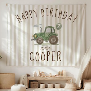 May include: A fabric wall hanging featuring a beige and white vertical striped background. The text HAPPY BIRTHDAY is printed in brown above a green tractor illustration with a green balloon showing the number 2. The name COOPER is below.