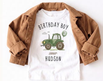 May include: White t-shirt with a green tractor graphic, the words "BIRTHDAY BOY" and "HUDSON" in black, and a green balloon with the number 3. The shirt is paired with a brown jacket.