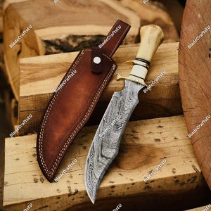 May include: A handcrafted hunting knife with a patterned blade and a bone handle, resting beside a brown leather sheath. The knife features a curved blade design and a brass bolster. The sheath is stitched with white thread.