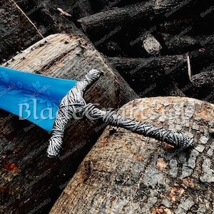 K&ouml;nnte beinhalten: Ein dekoratives Schwert mit einer blauen Klinge und einem zebragestreiften Griff liegt auf einem Haufen dunkelbrauner Baumst&auml;mme. Der Text "BladeCraftsEU" ist auf der Klinge zu sehen. Das Design des Schwertes deutet auf ein Fantasy- oder historisches Thema hin.