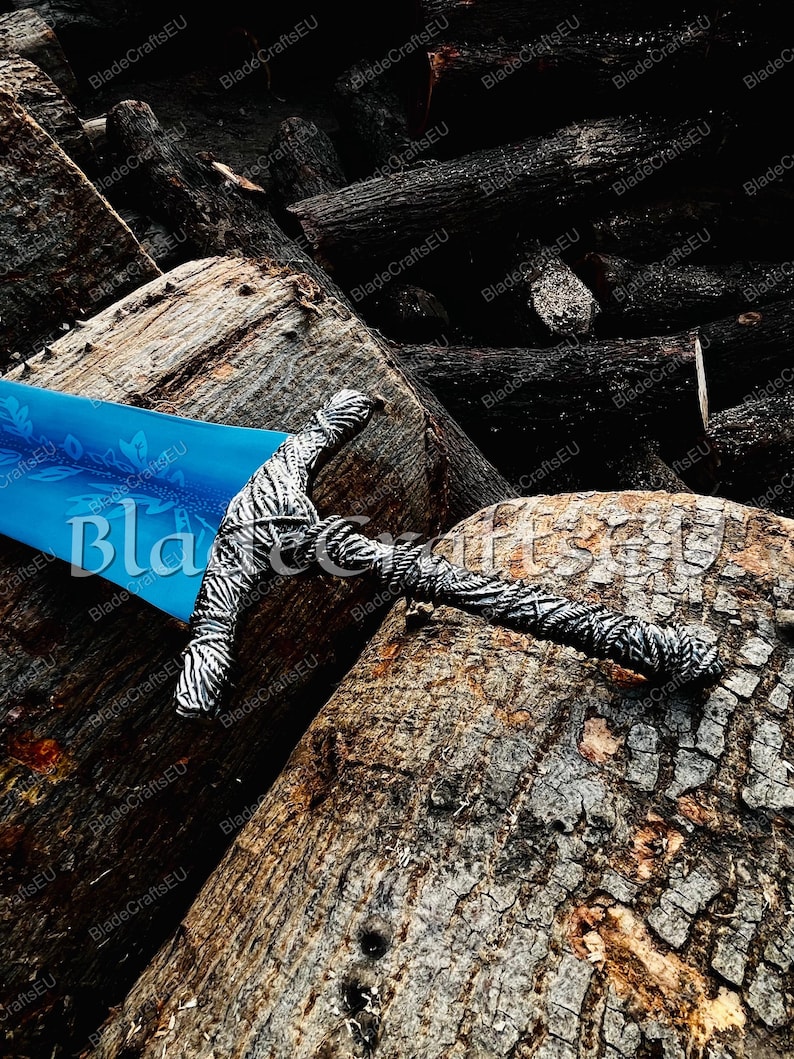 K&ouml;nnte beinhalten: Ein dekoratives Schwert mit einer blauen Klinge und einem zebragestreiften Griff liegt auf einem Haufen dunkelbrauner Baumst&auml;mme. Der Text "BladeCraftsEU" ist auf der Klinge zu sehen. Das Design des Schwertes deutet auf ein Fantasy- oder historisches Thema hin.