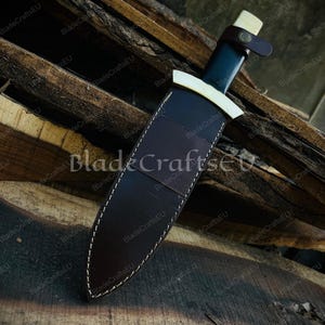 Puede incluir: Un cuchillo artesanal con una hoja oscura y un mango de color claro. El cuchillo est&aacute; en una funda de cuero marr&oacute;n con costuras blancas. Una correa de cuero asegura el cuchillo. El fondo es una superficie de madera.