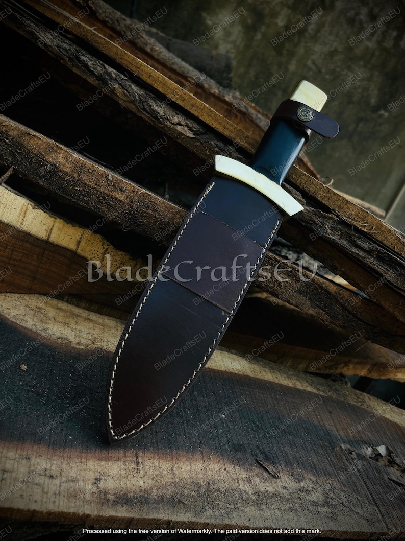 Puede incluir: Un cuchillo artesanal con una hoja oscura y un mango de color claro. El cuchillo est&aacute; en una funda de cuero marr&oacute;n con costuras blancas. Una correa de cuero asegura el cuchillo. El fondo es una superficie de madera.