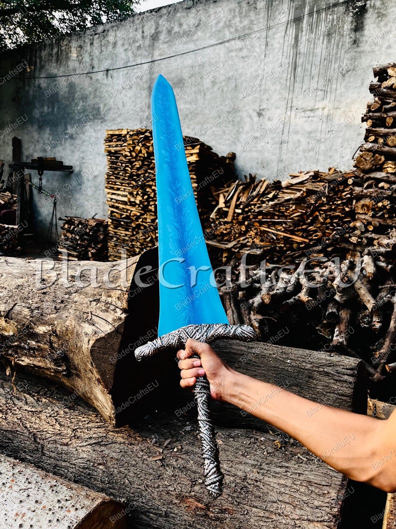 K&ouml;nnte beinhalten: Ein gro&szlig;es, blaues Schwert mit dekorativer Klinge und gemustertem Griff. Das Schwert wird vor einem Hintergrund aus Holz und Baumst&auml;mmen gehalten. Der Text "BladeCraftsEU" ist auf dem Bild sichtbar.
