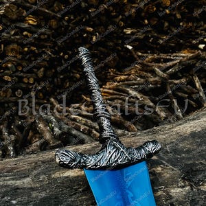 K&ouml;nnte beinhalten: Ein dekoratives Schwert mit einer blauen Klinge und einem verzierten silberfarbenen Knauf. Der Knauf weist aufwendige Details und einen strukturierten Griff auf. Die Klinge hat ein eingraviertes Blumendesign. Das Schwert wird auf einem Holzstamm ausgestellt.