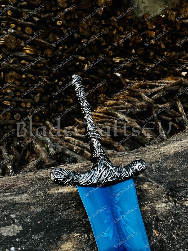 K&ouml;nnte beinhalten: Ein dekoratives Schwert mit einer blauen Klinge und einem verzierten silberfarbenen Knauf. Der Knauf weist aufwendige Details und einen strukturierten Griff auf. Die Klinge hat ein eingraviertes Blumendesign. Das Schwert wird auf einem Holzstamm ausgestellt.