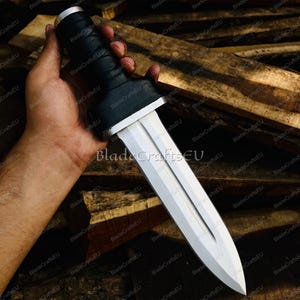 Puede incluir: Una espada gladius plateada y blanca con un mango negro. La hoja es de doble filo y tiene un canal central. El mango est&aacute; envuelto en material negro. La espada se sostiene en una mano sobre un fondo de madera.