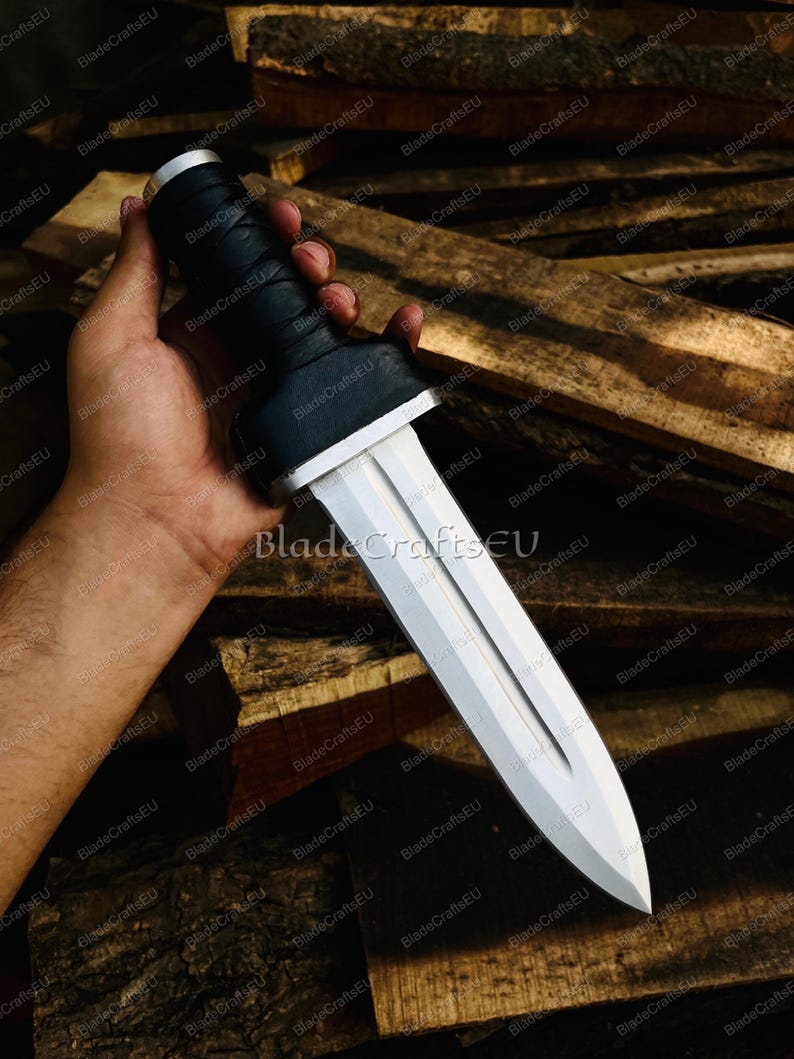 Puede incluir: Una espada gladius plateada y blanca con un mango negro. La hoja es de doble filo y tiene un canal central. El mango est&aacute; envuelto en material negro. La espada se sostiene en una mano sobre un fondo de madera.