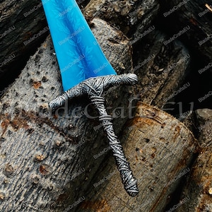 Pode incluir: Uma espada de fantasia com uma lâmina azul brilhante e uma empunhadura prateada detalhada. A lâmina tem um padrão decorativo. A espada repousa sobre uma pilha de toras de madeira, criando um cenário rústico.