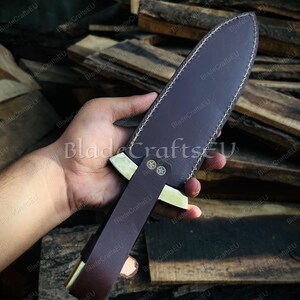 Puede incluir: Una funda de cuchillo de cuero marr&oacute;n con un mango decorativo de lat&oacute;n y una hoja. La funda est&aacute; cosida con hilo blanco. El cuchillo se sostiene en una mano sobre un fondo de tablones de madera.