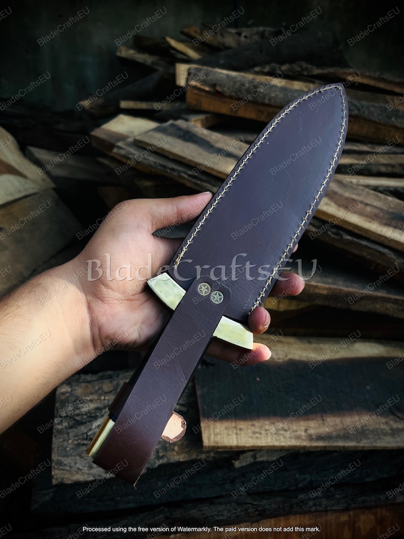 Puede incluir: Una funda de cuchillo de cuero marr&oacute;n con un mango decorativo de lat&oacute;n y una hoja. La funda est&aacute; cosida con hilo blanco. El cuchillo se sostiene en una mano sobre un fondo de tablones de madera.
