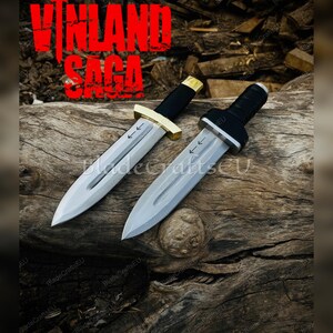 Puede incluir: Dos dagas plateadas con mangos negros y detalles dorados, expuestas sobre una superficie de madera envejecida. Las hojas tienen marcas de flechas. El texto "VINLAND SAGA" est&aacute; en rojo sobre las dagas.