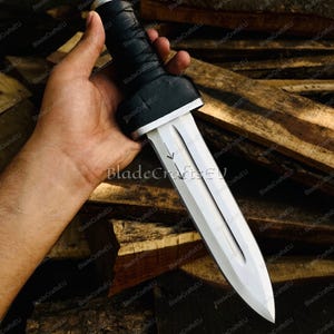 Puede incluir: Una daga plateada con una hoja de doble filo y un mango negro envuelto. La daga tiene un acento blanco en la base del mango. El texto "BladeCraftsEU" es visible en la hoja.