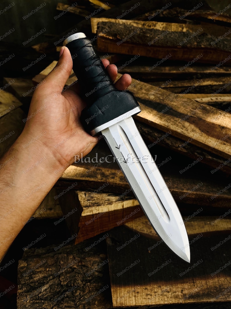 Puede incluir: Una daga plateada con una hoja de doble filo y un mango negro envuelto. La daga tiene un acento blanco en la base del mango. El texto "BladeCraftsEU" es visible en la hoja.