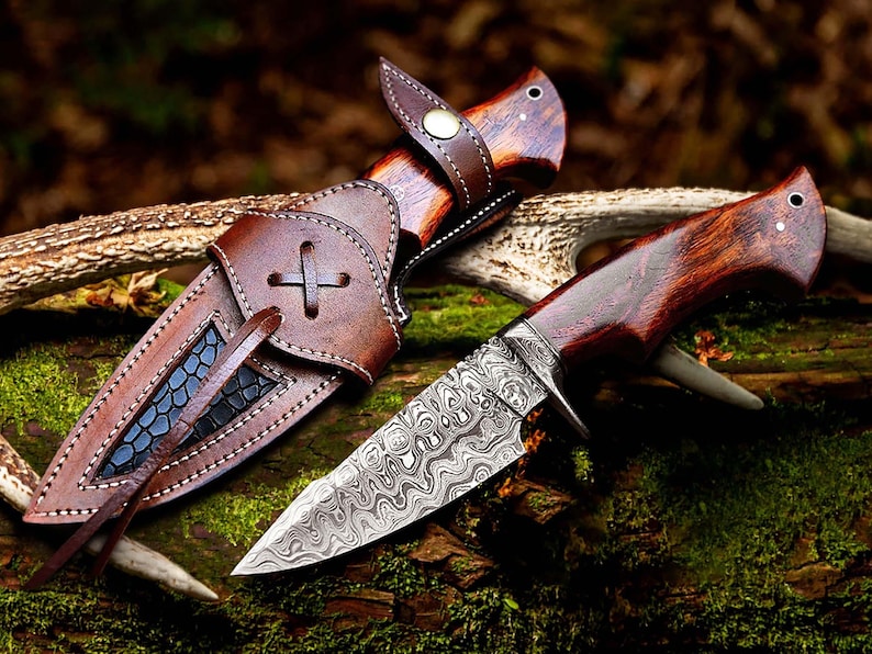Puede incluir: Un cuchillo de caza artesanal con una hoja de acero estampada y un mango de madera. El cuchillo est&aacute; junto a su funda de cuero marr&oacute;n, que tiene un dise&ntilde;o de cruz decorativa. El cuchillo y la funda se exhiben sobre una superficie natural y musgosa.