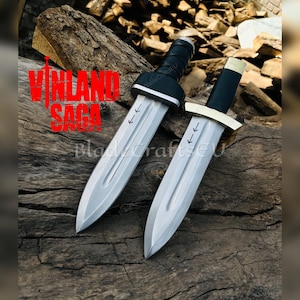 Puede incluir: Dos dagas plateadas con mangos negros y el texto "VINLAND SAGA" en rojo. Las dagas se exhiben sobre una superficie de madera. Cada daga tiene una hoja plateada y un mango negro.