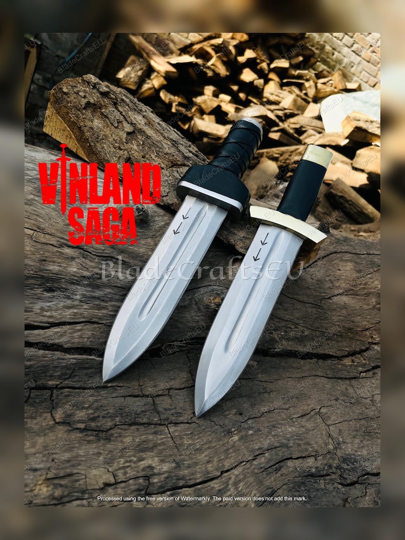 Puede incluir: Dos dagas plateadas con mangos negros y el texto "VINLAND SAGA" en rojo. Las dagas se exhiben sobre una superficie de madera. Cada daga tiene una hoja plateada y un mango negro.