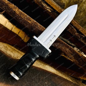 Puede incluir: Una daga plateada con una hoja puntiaguda y un mango envuelto en cuero negro. La hoja presenta un dise&ntilde;o decorativo de flecha. La daga se muestra sobre un fondo de tablones de madera, destacando su dise&ntilde;o.