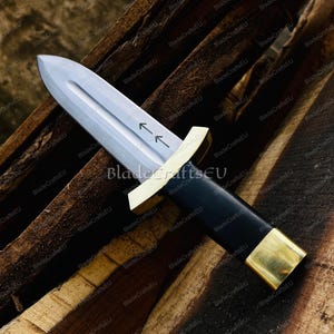 Puede incluir: Una daga con una hoja plateada y un mango negro. El mango est&aacute; acentuado con metal dorado en la guarda y el pomo. La hoja tiene dos flechas grabadas. La daga descansa sobre una superficie de madera.