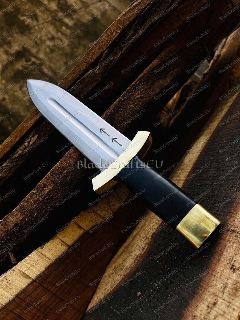Puede incluir: Una daga con una hoja plateada y un mango negro. El mango est&aacute; acentuado con metal dorado en la guarda y el pomo. La hoja tiene dos flechas grabadas. La daga descansa sobre una superficie de madera.