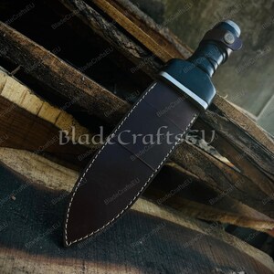 Puede incluir: Una espada romana gladius con una funda de cuero marr&oacute;n oscuro. La espada tiene un mango negro y una guarda plateada. La funda est&aacute; cosida con hilo blanco. La hoja no es visible. Sobre tablas de madera.