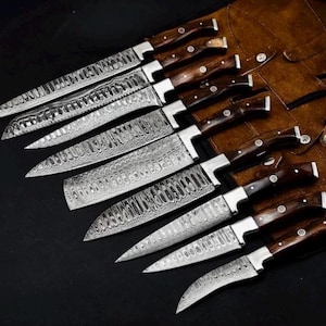Op de afbeelding: Een set van acht Damascus stalen keukenmessen met donkerhouten handvatten, gerangschikt op een donkere ondergrond. De messen hebben een uniek patroon. Een bruine leren messenrol is zichtbaar.
