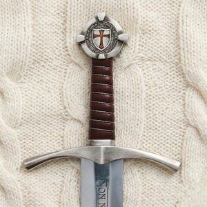 Peut inclure: Épée de style médiéval avec une lame argentée et une poignée enveloppée de cuir marron. La garde présente une croix argentée avec une croix rouge au centre. L'épée est posée sur un fond blanc texturé.