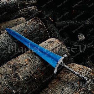 K&ouml;nnte beinhalten: Ein blaues Schwert mit einem detaillierten silbernen Griff liegt auf einem Haufen dunkelbrauner Baumst&auml;mme. Die Klinge hat ein strukturiertes Aussehen, und der Griff weist komplizierte Muster auf. Das Schwert ist ein Fantasy-Themenartikel.