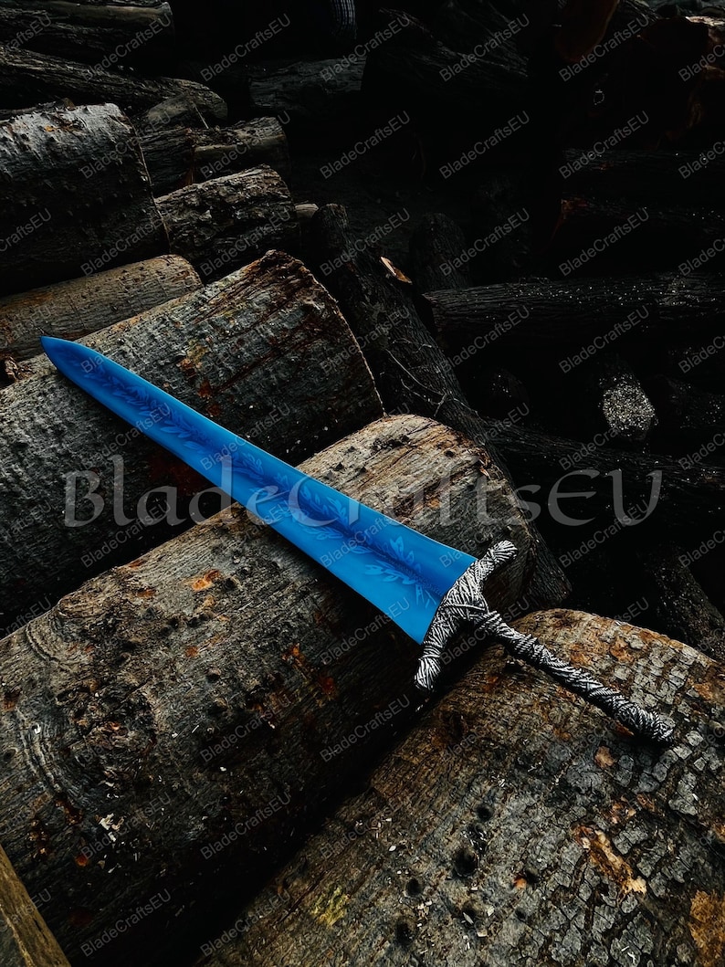 K&ouml;nnte beinhalten: Ein blaues Schwert mit einem detaillierten silbernen Griff liegt auf einem Haufen dunkelbrauner Baumst&auml;mme. Die Klinge hat ein strukturiertes Aussehen, und der Griff weist komplizierte Muster auf. Das Schwert ist ein Fantasy-Themenartikel.