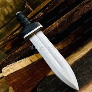 Puede incluir: Una espada plateada con empu&ntilde;adura negra y guarda blanca. La hoja de doble filo termina en punta. La espada se exhibe sobre una superficie de madera. El texto "BladeCraftsEU" es visible.