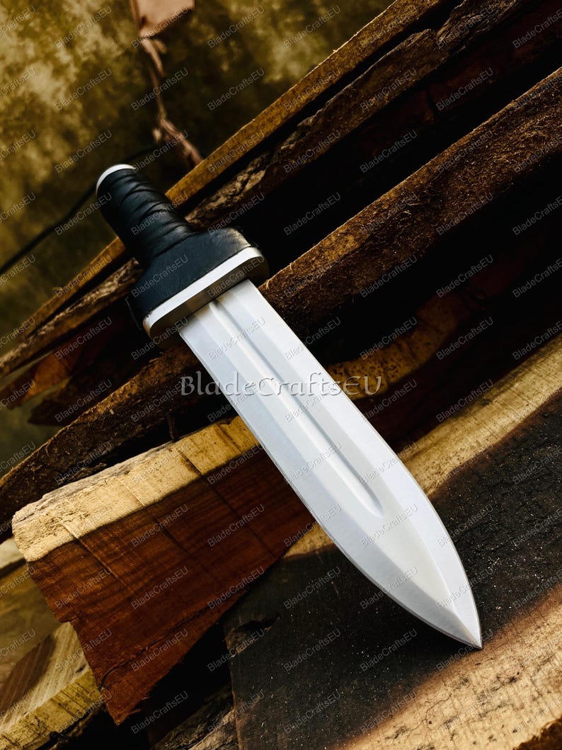 Puede incluir: Una espada plateada con empu&ntilde;adura negra y guarda blanca. La hoja de doble filo termina en punta. La espada se exhibe sobre una superficie de madera. El texto "BladeCraftsEU" es visible.