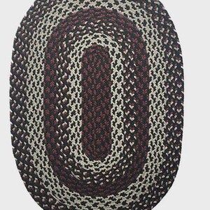 May include: Oval braided rug in shades of burgundy, cream, and black. The rug features a central burgundy oval surrounded by cream and black braided sections. A decorative floor covering for any room.