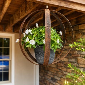 May include: A hanging planter featuring a dark gray textured pot nestled inside a spherical frame made of three intersecting, weathered, rust-colored metal bands. A lush green plant with small white flowers grows from the central pot.