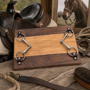 May include: A rectangular wooden board featuring a central light oak panel flanked by two darker walnut wood sections. Two silver-toned metal horse snaffle bits are attached to the board with small brown leather straps and silver rivets.