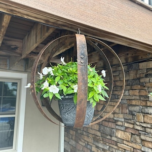May include: Hanging metal sphere planter with a dark gray pot filled with green foliage and white flowers. The planter is suspended from a wooden structure and set against a brick wall. The metal has a rustic, weathered appearance.