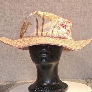 May include: A wide-brimmed hat with a patterned crown and brim. The crown features a beige fabric with brown leaf designs and red floral accents. The brim is woven with a light brown, textured material. The hat is displayed on a black mannequin head.