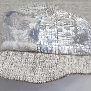 May include: A beige bucket hat with a wide brim. The hat features a patchwork design on the crown with various textured fabrics in shades of gray, blue, and white. The brim is a solid beige color.