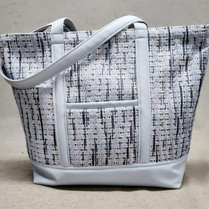 May include: A light blue tote bag with a woven pattern in white and navy blue. The bag has a light blue base, handles, and trim. A small pocket is visible on the front of the bag.