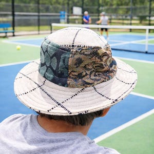 May include: A cream-colored bucket hat with a black grid pattern and patterned fabric patches in shades of green and brown. The hat is worn by a person on a tennis court.