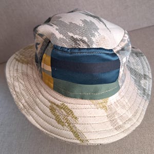 May include: A patchwork bucket hat with a wide brim. The hat features a mix of patterns and colors, including blue, green, yellow, and gray. The brim has a light green leaf-like pattern. The crown has a quilted appearance.