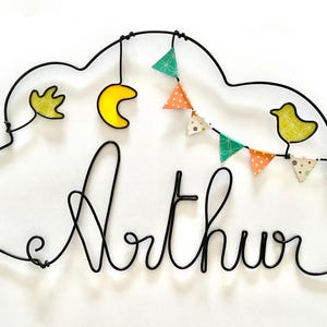 May include: A black wire cloud-shaped wall decoration with the name "Arthur" in cursive. It features a yellow crescent moon, a yellow bird, and a string of colourful pennant flags. The cloud is adorned with a yellow leaf.