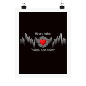 May include: A black poster with the text "Heart rate: f/stop perfection." The design features a red heart in the center, surrounded by a camera aperture graphic. A white waveform graphic spans the top and bottom of the heart.