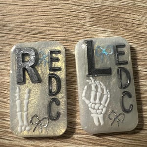 Puede incluir: Dos piezas rectangulares de resina con las letras "RED C" y "LED C" en gris oscuro. Una presenta una mano de esqueleto, la otra una mano de esqueleto y huesos. Ambas tienen lazos plateados y un fondo claro.